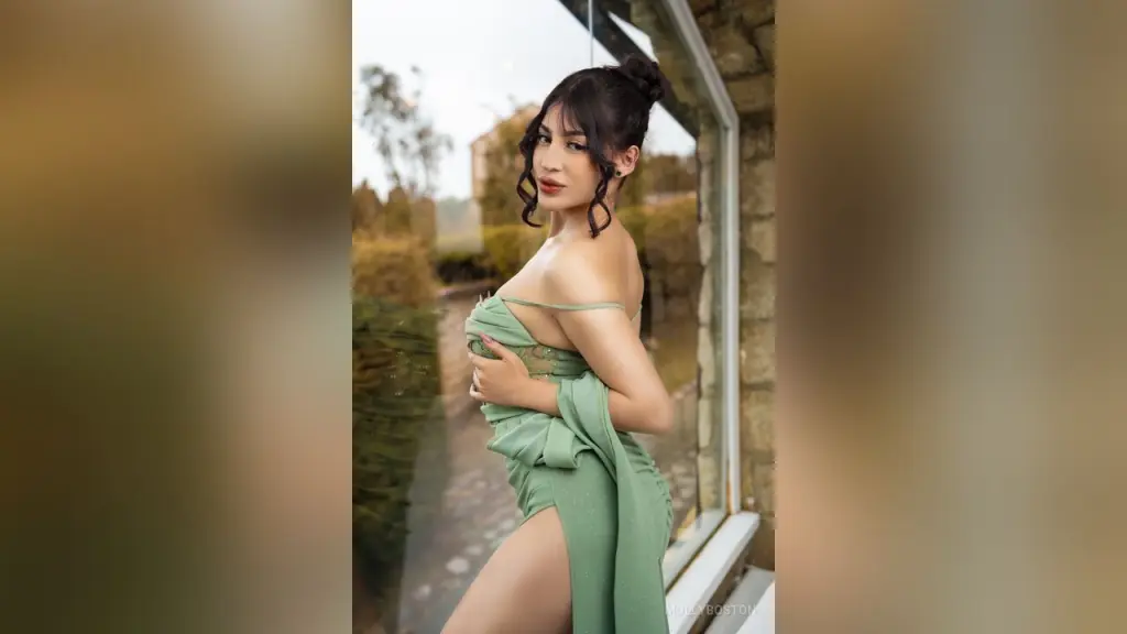 MollyBoston — MollyBoston poses by a window wearing a low-cut, sage green dress with a high slit.