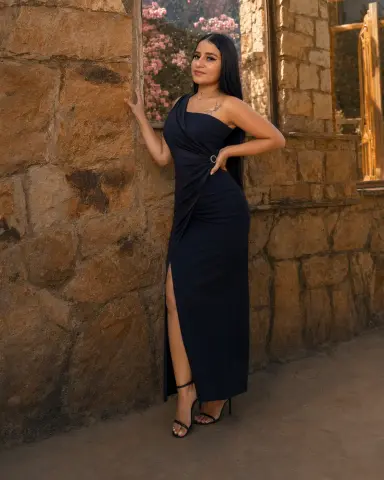 Elegant woman in a navy blue evening gown posing by a rustic stone wall in warm sunlight.