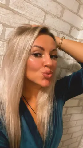 Blonde woman in a teal velvet top making a playful kissy face against a white brick wall.