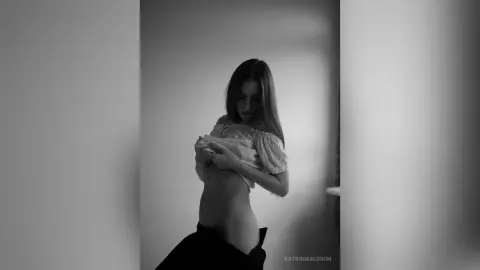 Moody black and white portrait of a woman in a white off-the-shoulder blouse posing in a minimalist room.
