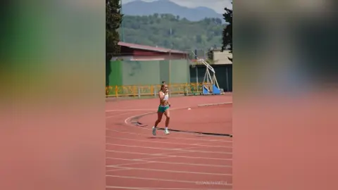 Athletic woman in green sports bra and shorts running on a red track field under a bright sky.