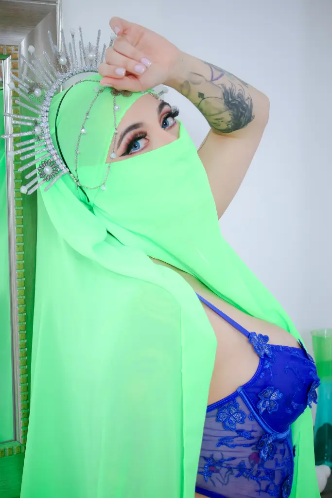 Sashaarnab poses with her hands near her face, wearing a black head covering and a red top against a dark background.