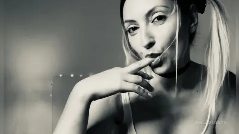 Blonde woman in a white tank top posing with a thoughtful expression in a moody black and white portrait.