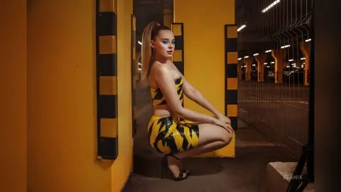 BeaNix squats in a yellow and black patterned tube top and skirt set against a dark industrial background.