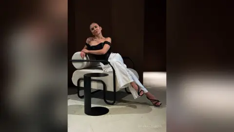 MilanaVolkova sits on a white chair wearing a black off-the-shoulder top and white pants, posing with her legs crossed.