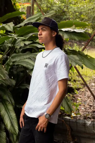 KylePresly stands outdoors wearing a white t-shirt and black pants, looking off to the side near lush green tropical plants.