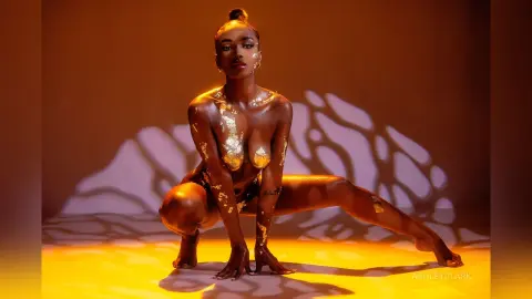 Glamorous woman with gold body paint posing in a dramatic sunlit studio setting.