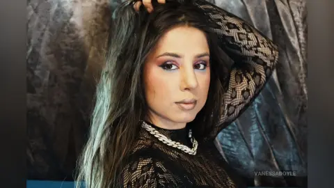 Glamorous woman in a black lace top with dramatic makeup posing against a textured grey backdrop.