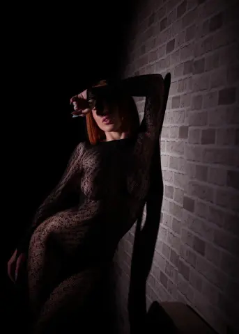 Redheaded woman in a sheer black patterned bodysuit posing against a brick wall in moody low light.