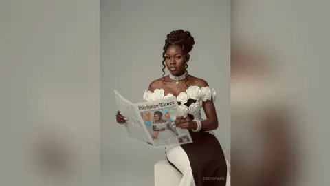 ZoeySpark poses in a white off-the-shoulder floral dress, holding a newspaper titled Birthday Times against a plain grey background.