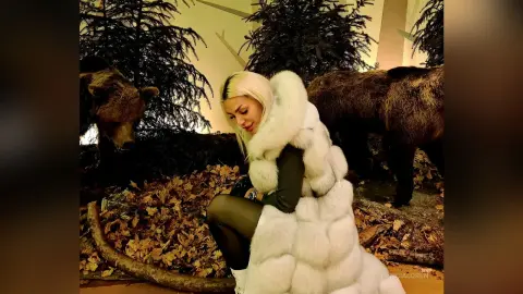 Blonde woman in a white faux fur coat sitting amidst autumn leaves in a rustic woodland setting.