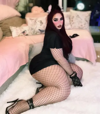 Glamorous woman in a black dress and fishnet tights sitting on a white rug in a cozy pink bedroom.