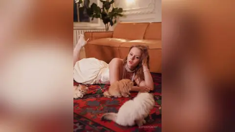 SophiePeachy lies on a patterned rug in a white dress, surrounded by several small fluffy cats in a living room.