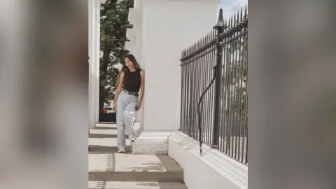 LaurentBelluci stands outdoors in a black tank top and light blue jeans against a white wall and black metal fence.