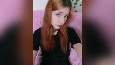Redheaded woman in a black t-shirt and choker sitting against a soft pink background with a calm expression.