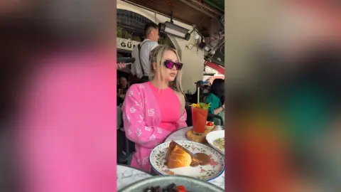 Blonde woman in a pink outfit enjoying a summer brunch with cake and drinks at an outdoor cafe.