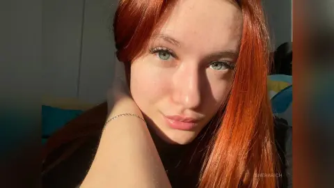 SheraRich looks into the camera with her long red hair and light eyes, resting her hand against her neck.
