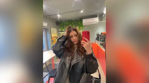 Young woman in a black leather jacket taking a mirror selfie in a modern cafe setting.