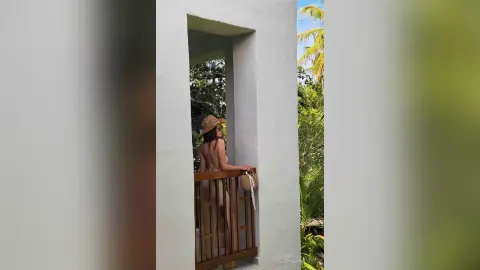Woman in a tan summer outfit and straw hat relaxing on a tropical villa balcony overlooking lush palm trees.
