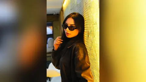 Stylish woman in black sunglasses and leather jacket leaning against a textured gold wall in warm lighting.