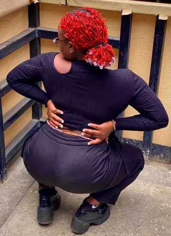 PrettyDoll22 poses with her back to the camera, wearing a black long-sleeve top and black pants, showing her red hair and red-tipped braids.