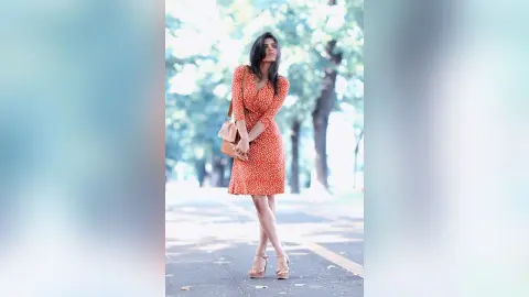 Stylish woman in a red polka dot summer dress standing on a sunlit tree-lined street.