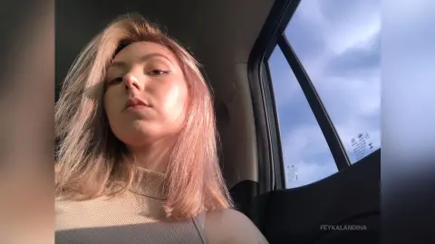 Young woman with blonde hair in a beige ribbed top sitting in a car during golden hour sunset.
