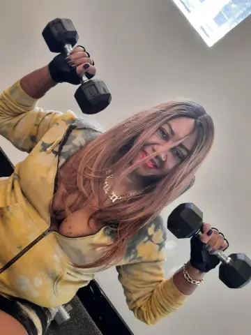 Smiling woman in yellow hoodie lifting black dumbbells during an intense home gym workout session.