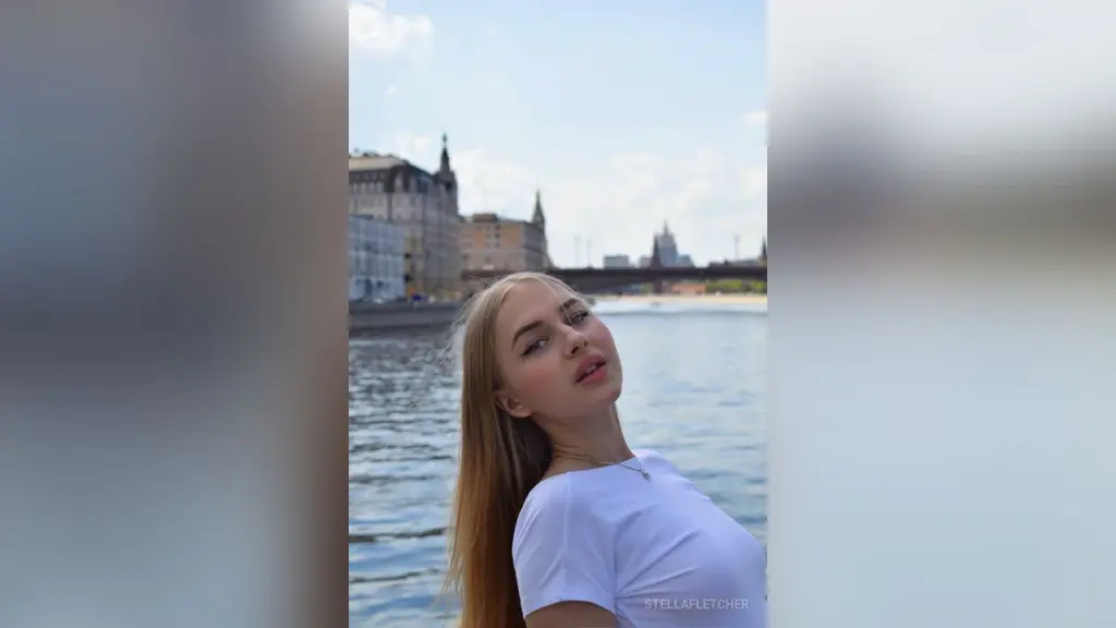 StellaFletcher โ StellaFletcher tilts her head back while wearing a white t-shirt against a river and city skyline background.