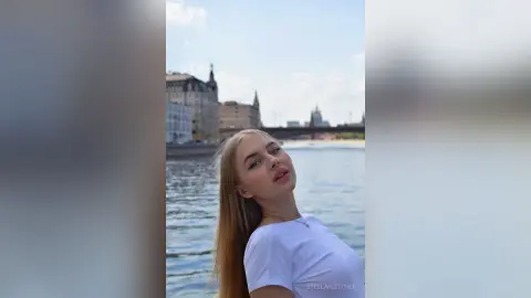 StellaFletcher tilts her head back while wearing a white t-shirt against a river and city skyline background.