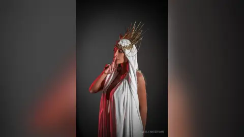 LeslyAnderson wears a white flowing gown and a gold spiked crown against a dark grey background.