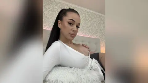 AriaVex poses in a white long-sleeve top while leaning against a white furry pillow.