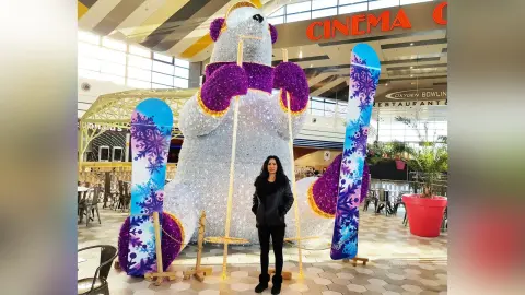 Woman in black jacket standing by a giant glittery polar bear sculpture and blue skis in a modern mall setting.
