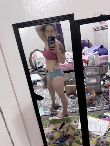 Young woman with tattoos in a pink crop top and denim shorts posing in a messy bedroom mirror selfie.