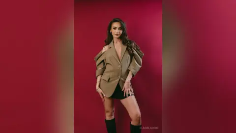 Confident woman in a stylish tan cutout blazer and black shorts posing against a deep red studio background.