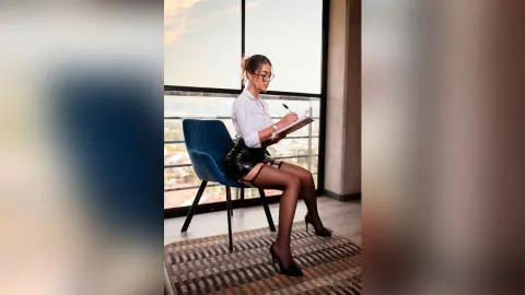 Professional woman in white blouse and black leather skirt writing in a notebook by a bright office window.