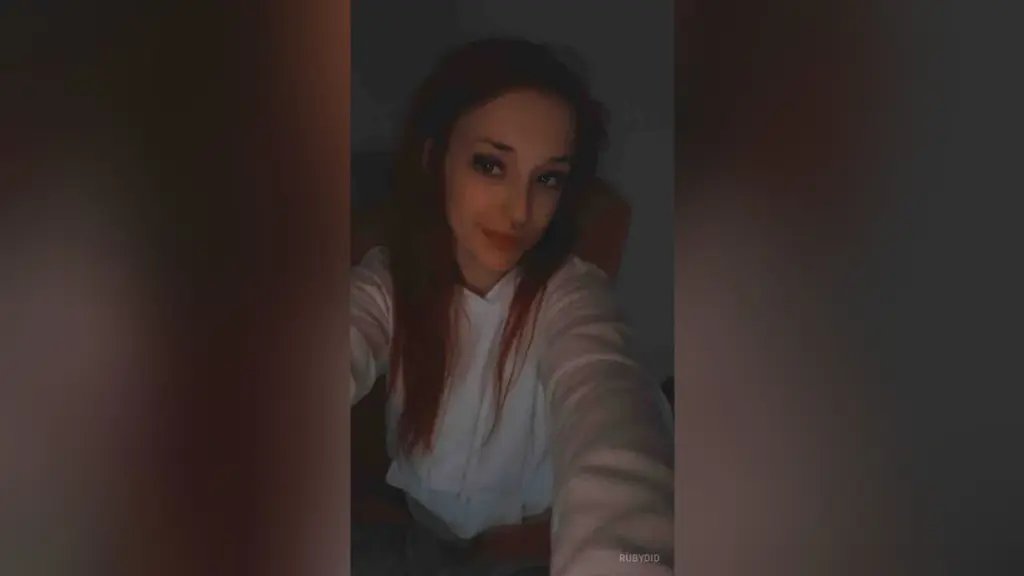 Red-haired woman with nose ring taking a selfie in a white tank top using a sparkly rhinestone phone case.