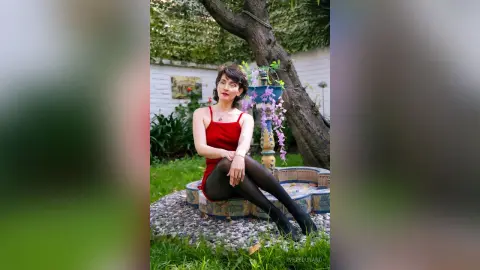 IveteDurand wears a red tank top and black tights while sitting on a stone fountain edge in a garden setting.