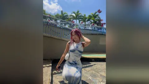 MilaGreco poses outdoors in a white and blue tie-dye dress with her red hair down, standing on a stone patio near a blue railing and palm trees.