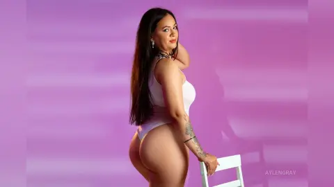Curvy woman in a white bodysuit posing against a purple background for a glamorous studio photoshoot.
