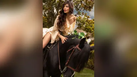 SophiaNapoles with long wavy hair, wearing a light yellow dress, sitting on a black horse in an outdoor setting.
