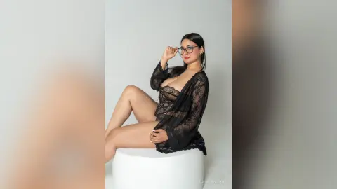 Glamorous woman in a black lace robe sitting on a white stool against a minimalist studio background.