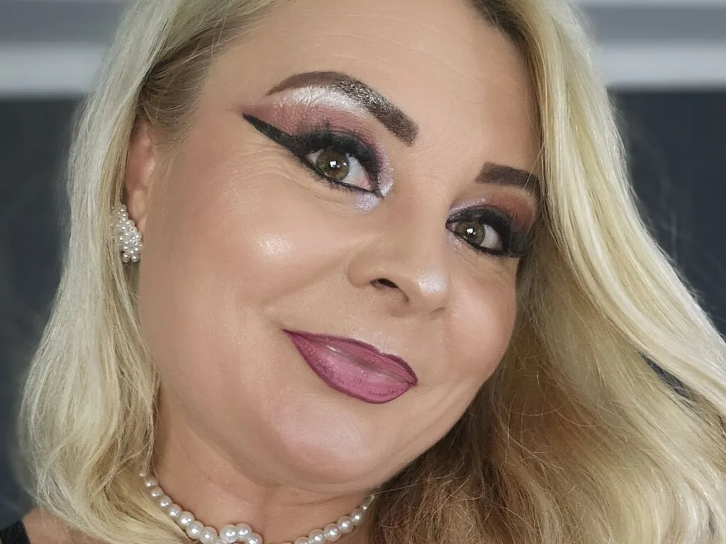 CatherineMill smiles at the camera wearing sparkly eye makeup, pink lipstick, and a pearl necklace.