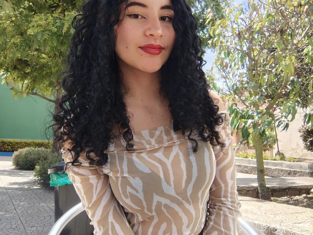 Young woman with curly dark hair wearing a beige zebra print top in a sunny outdoor garden setting.