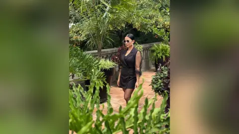 JuliahLewis stands outdoors in a black sleeveless dress surrounded by lush green plants and trees.
