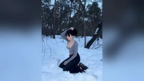 MarieJosePhilip poses in a snowy forest, wearing a tight grey long-sleeve top and black pants while kneeling in the snow with her hands in her dark hair.