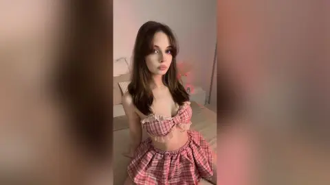 Young woman in a pink plaid crop top and matching skirt posing in a cozy, dimly lit bedroom.