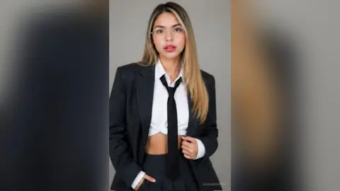 Young woman in a professional black suit and white shirt with a black tie posing against a neutral background.
