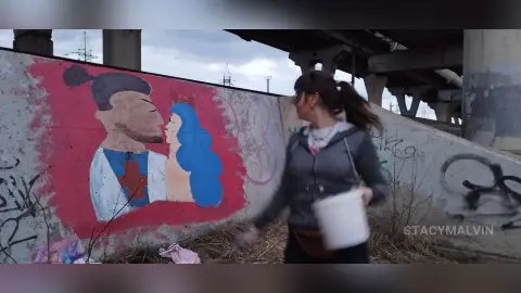 StacyMalvin stands outdoors near a concrete wall with a red mural, wearing a grey zip-up hoodie and holding a white bucket.