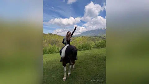 EvelynMills sits upright on a white and black horse in a grassy field under a blue sky.